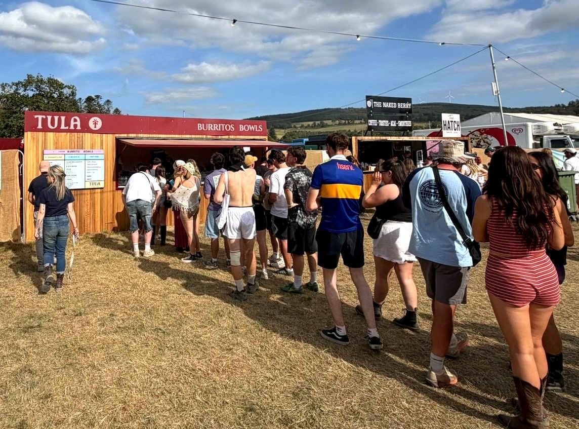 A long queue in the sun at the Tula trailer at a festival, with The Naked Berry sign behind