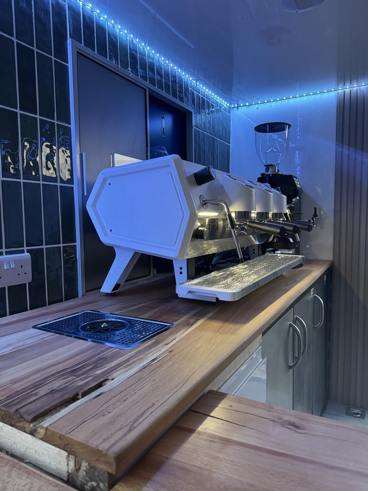 A Sanremo three-group espresso machine and grinder on a solid timber counter, with a flush-fitted knock box and LED-lit green tiles
