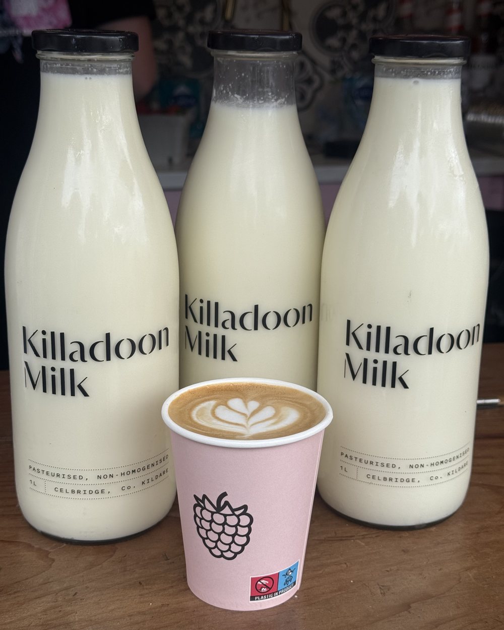 A latte with latte art in a pink Naked Berry cup in front of three glass bottles of Killadoon Milk